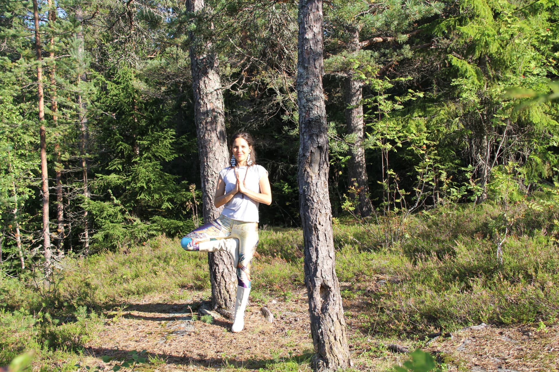 Karina standing in tree pose, hands in prayer, rooted in a sunlit pine forest, connected with Earth